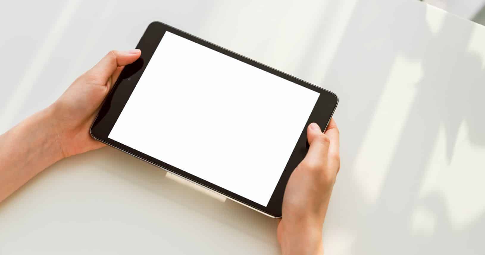 Mockup einer Hand, die ein digitales Tablet mit leerem Bildschirm auf einem Schreibtisch im Büro hält. Nutzen Sie Ihren Bildschirm für Werbezwecke.